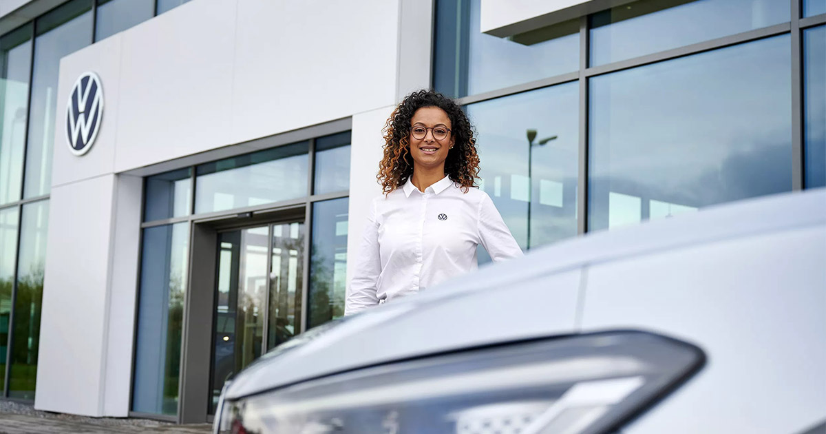 Woman in white shirt standing in front of Volkswagen dealership with a white car, showcasing professional automotive service.